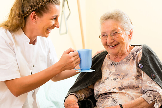 A care home nurse giving a smiling older lady a cup of tea while in a care home